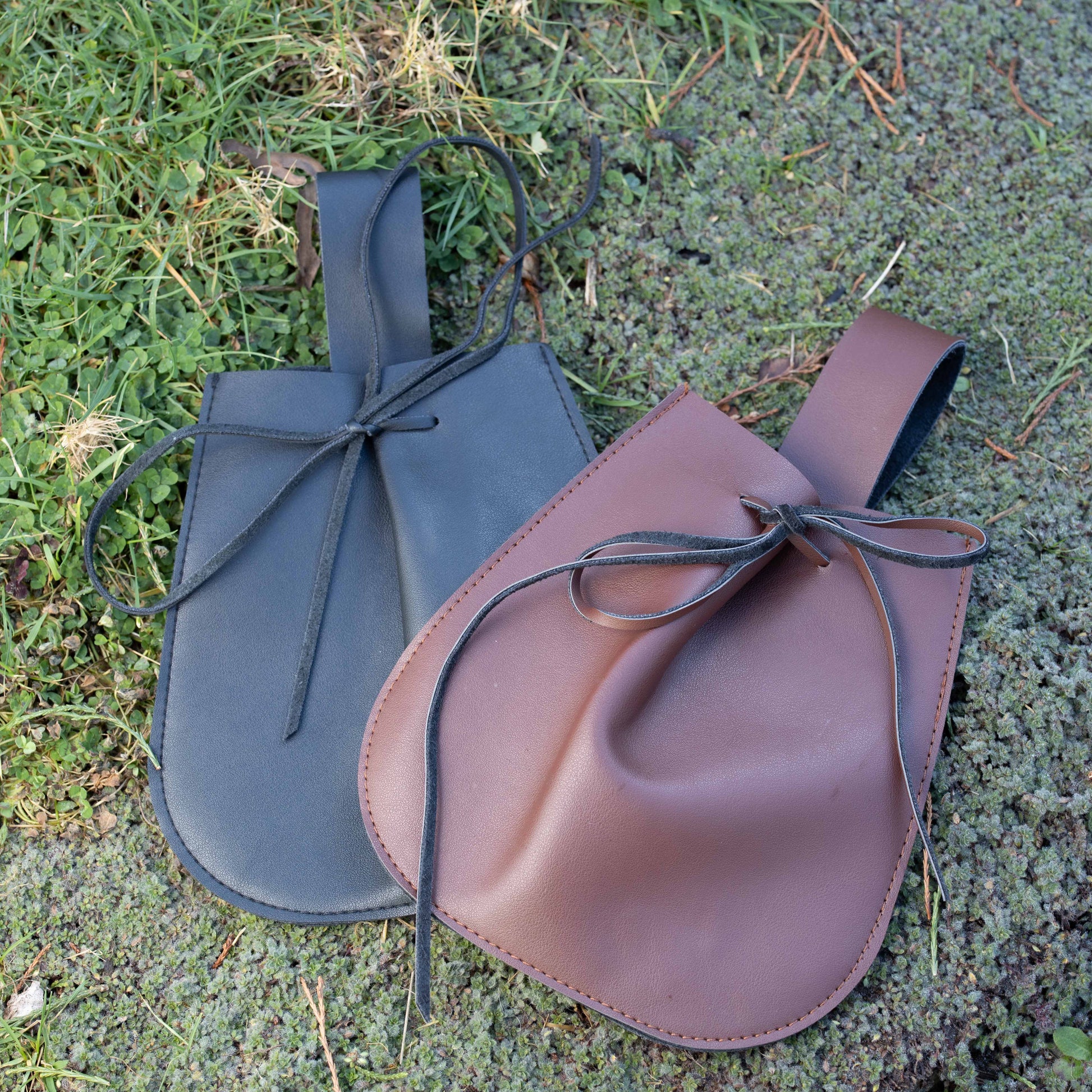 Two vegan leather drawstring pouches in brown and black designed to attach to belts up to 3 inches wide outdoors on grass.