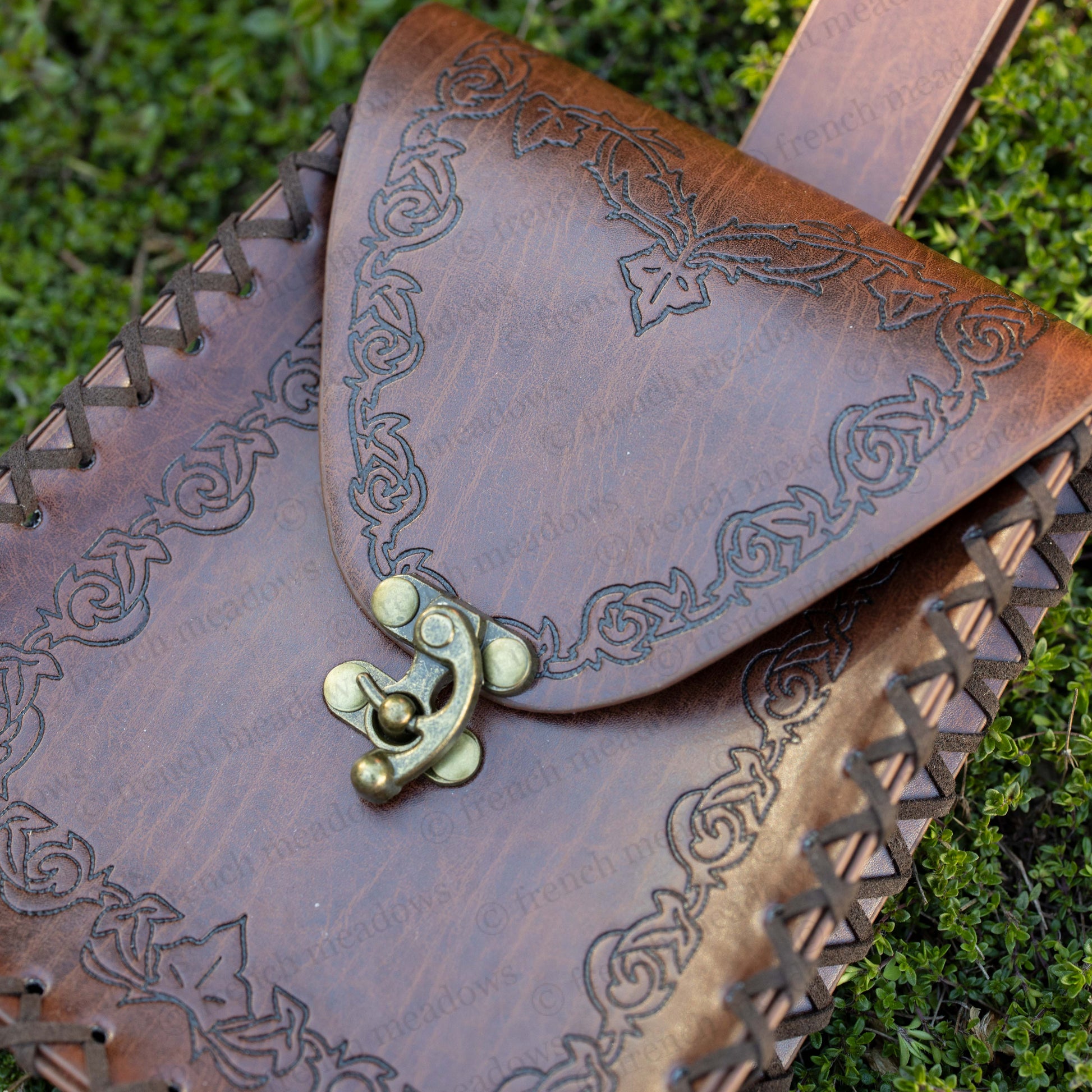 Elven Medieval Belt Pouch with brown leather, intricate scroll design, brass clasp, and woven edges on grass background