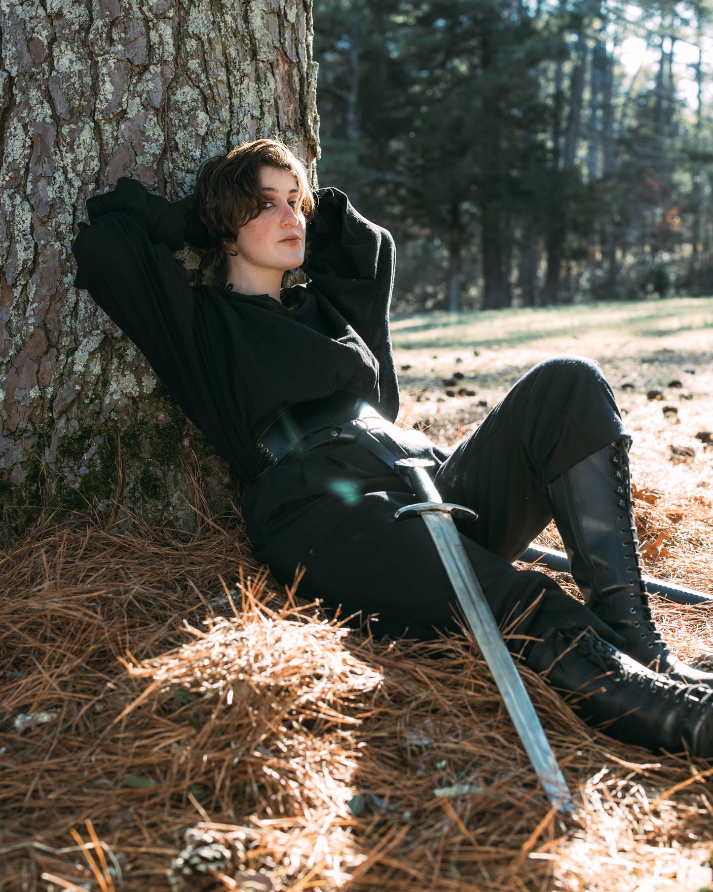 Person in black boots and pants leaning against a tree holding a sword, wearing Darcy Shirt in outdoor setting