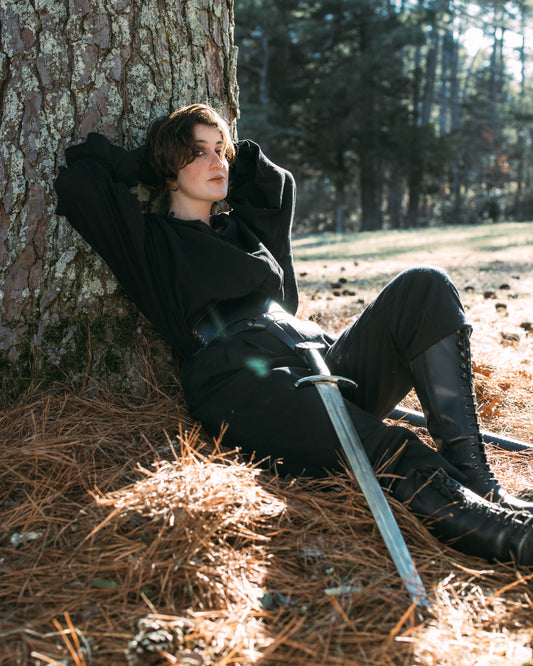 Person in black boots and pants leaning against a tree holding a sword, wearing Darcy Shirt in outdoor setting