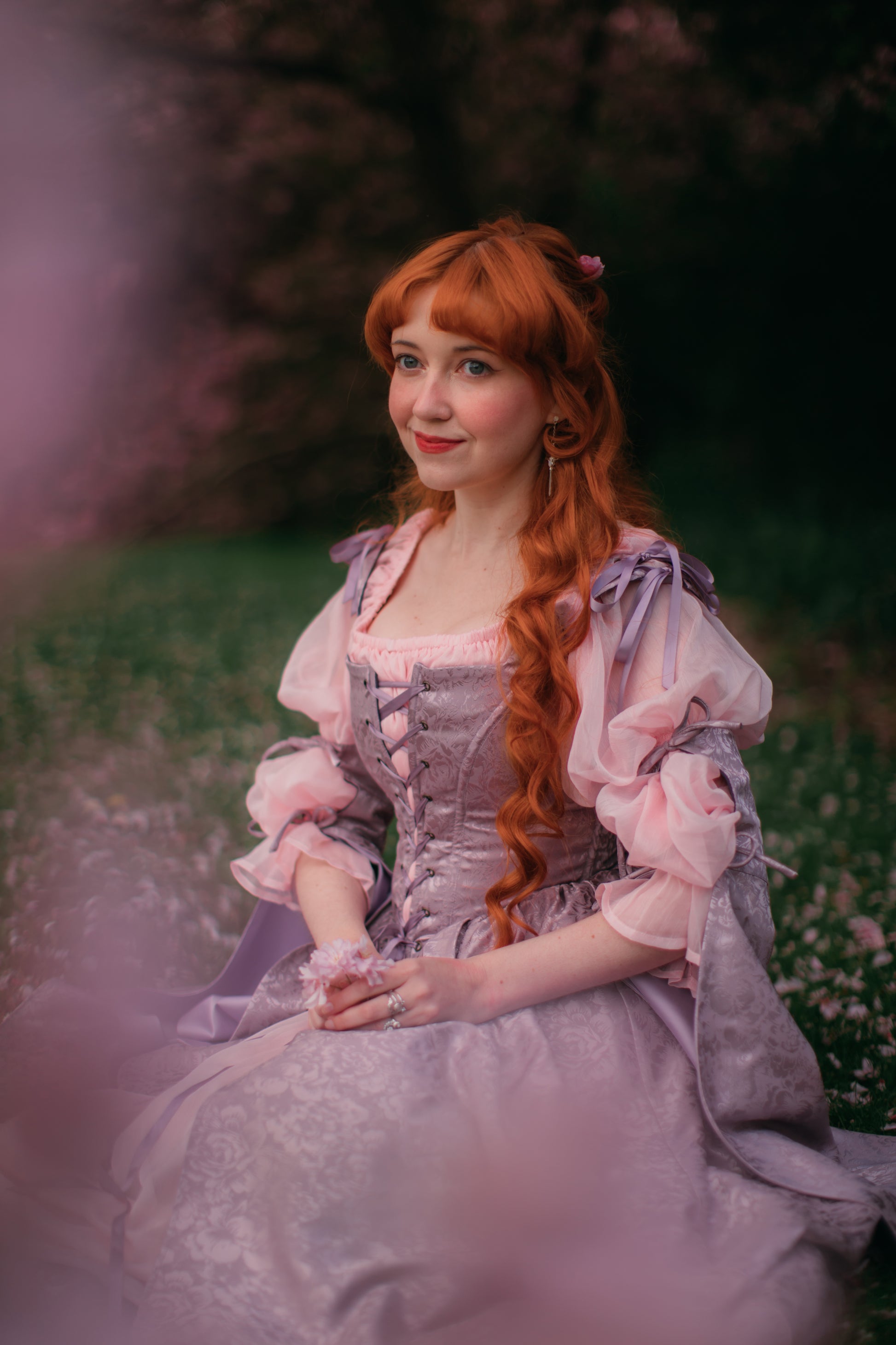 Young woman with red hair wearing lavender floral satin corset dress seated outdoors in garden setting.
