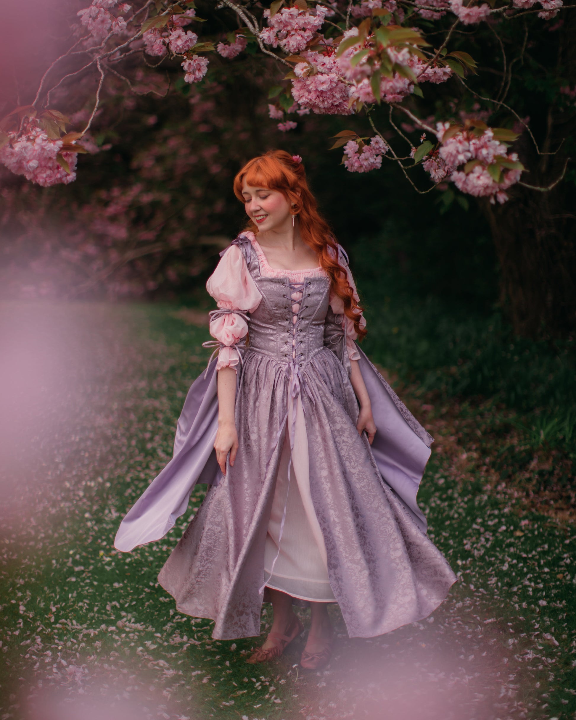 Lavender Floral Satin Corset Dress worn in a garden with blooming trees, featuring adjustable lacing and delicate bow straps.