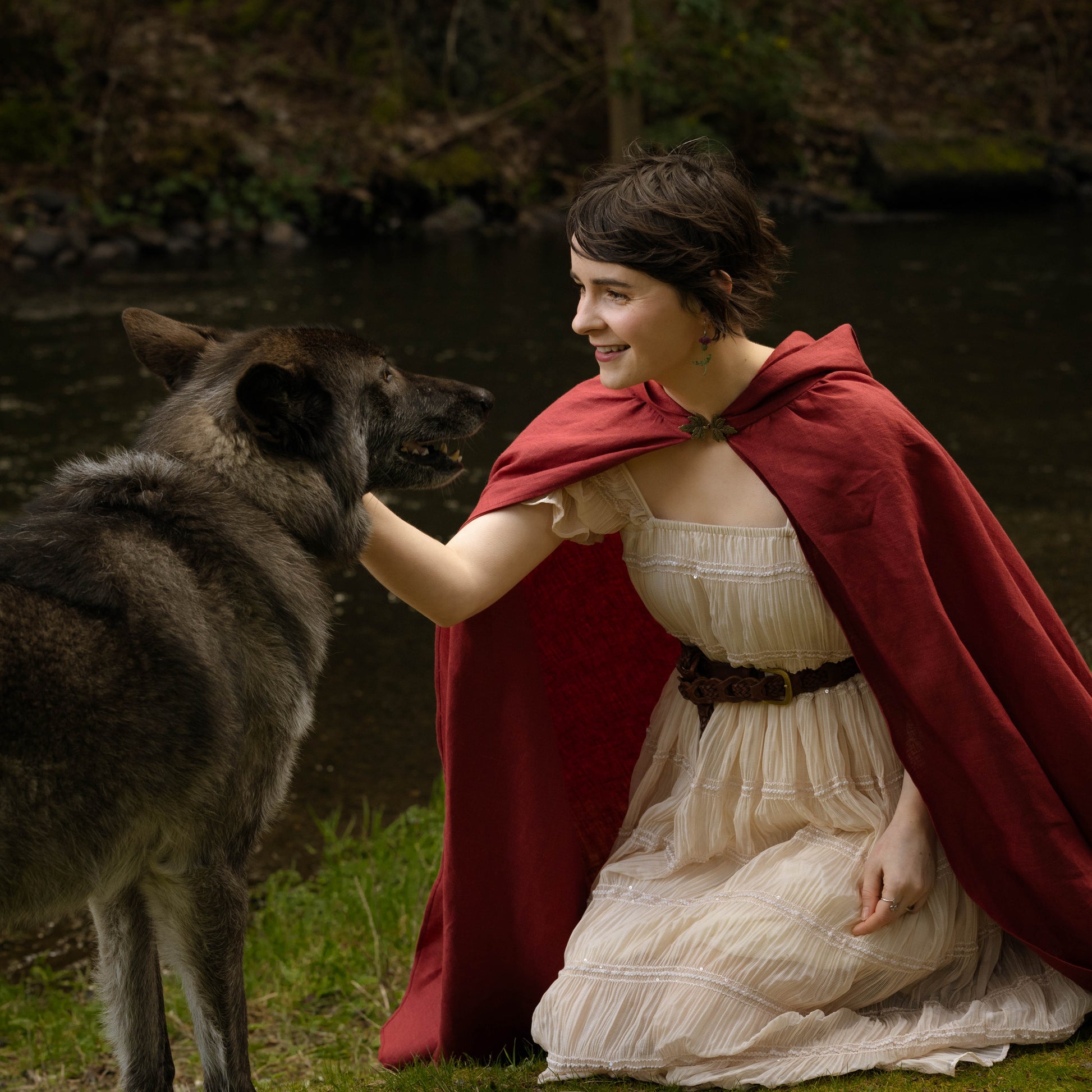 Woman wearing a red linen renaissance cloak kneeling by a river gently petting a large wolf-like dog.