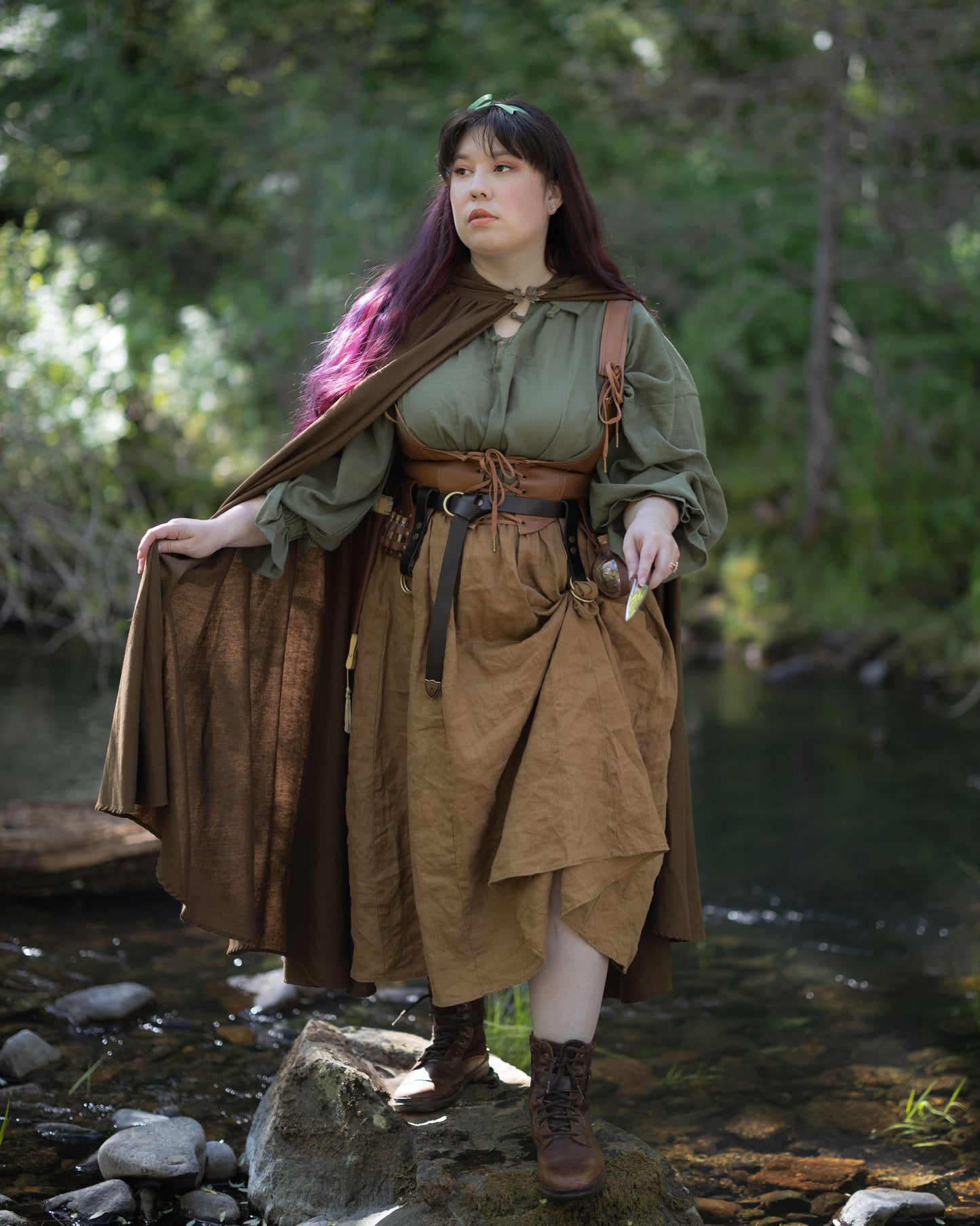 Woman wearing Linen Renaissance Cloak in brown, standing on rock by forest stream in renaissance-style outfit
