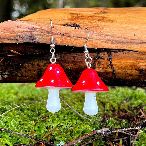Amanita Mushroom Earrings featuring bright red caps with white spots on white stems, displayed on wooden log over moss