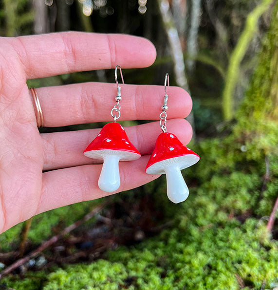 Amanita Mushroom Earrings featuring bright red caps with white spots on white stems held against mossy natural background