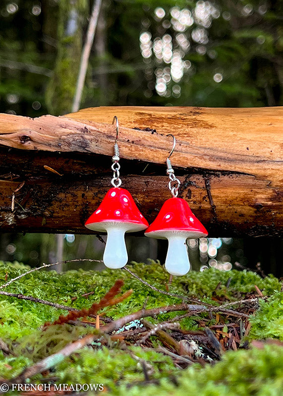Amanita Mushroom Earrings displayed on rustic wood against mossy forest background, featuring red caps with white spots and stems
