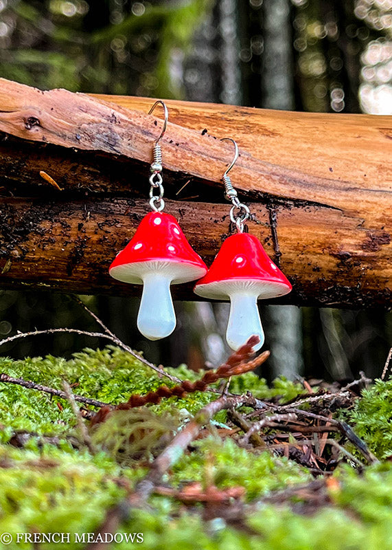 Amanita Mushroom Earrings displayed on wooden log with mossy forest background, featuring red caps with white spots and stems