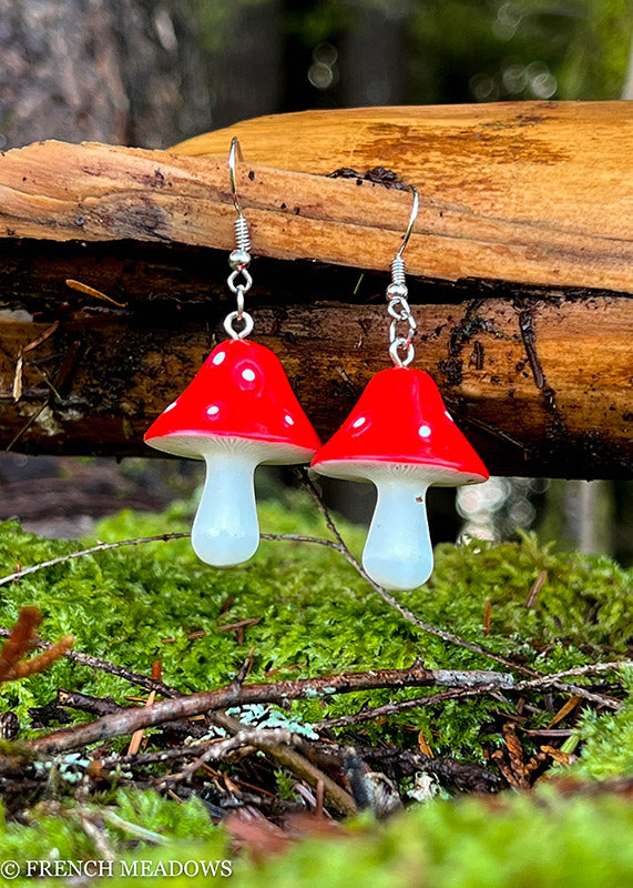 Amanita Mushroom Earrings featuring red and white spotted toadstool charms displayed on mossy woodland setting with natural log backdrop