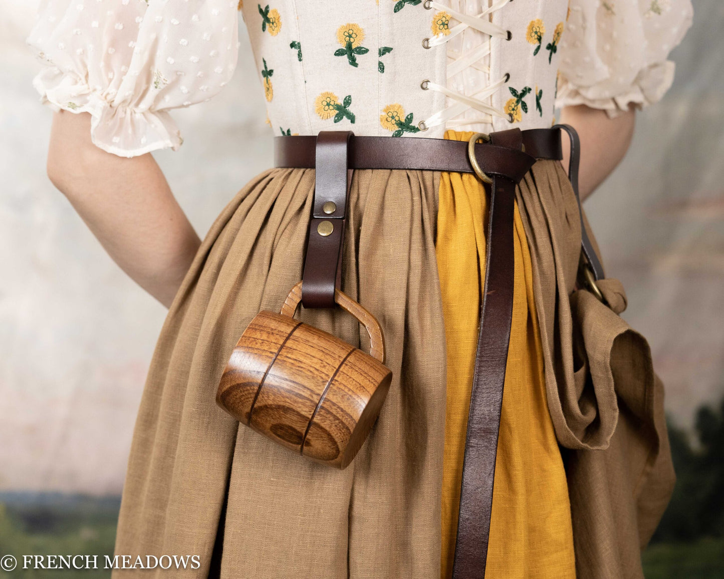 Brown leather tankard strap attached to belt, holding wooden mug against tan and yellow gathered skirt with floral print top
