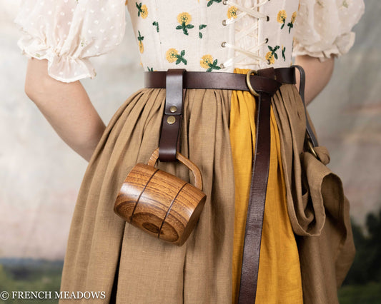 Brown leather tankard strap attached to belt, holding wooden mug against tan and yellow gathered skirt with floral print top