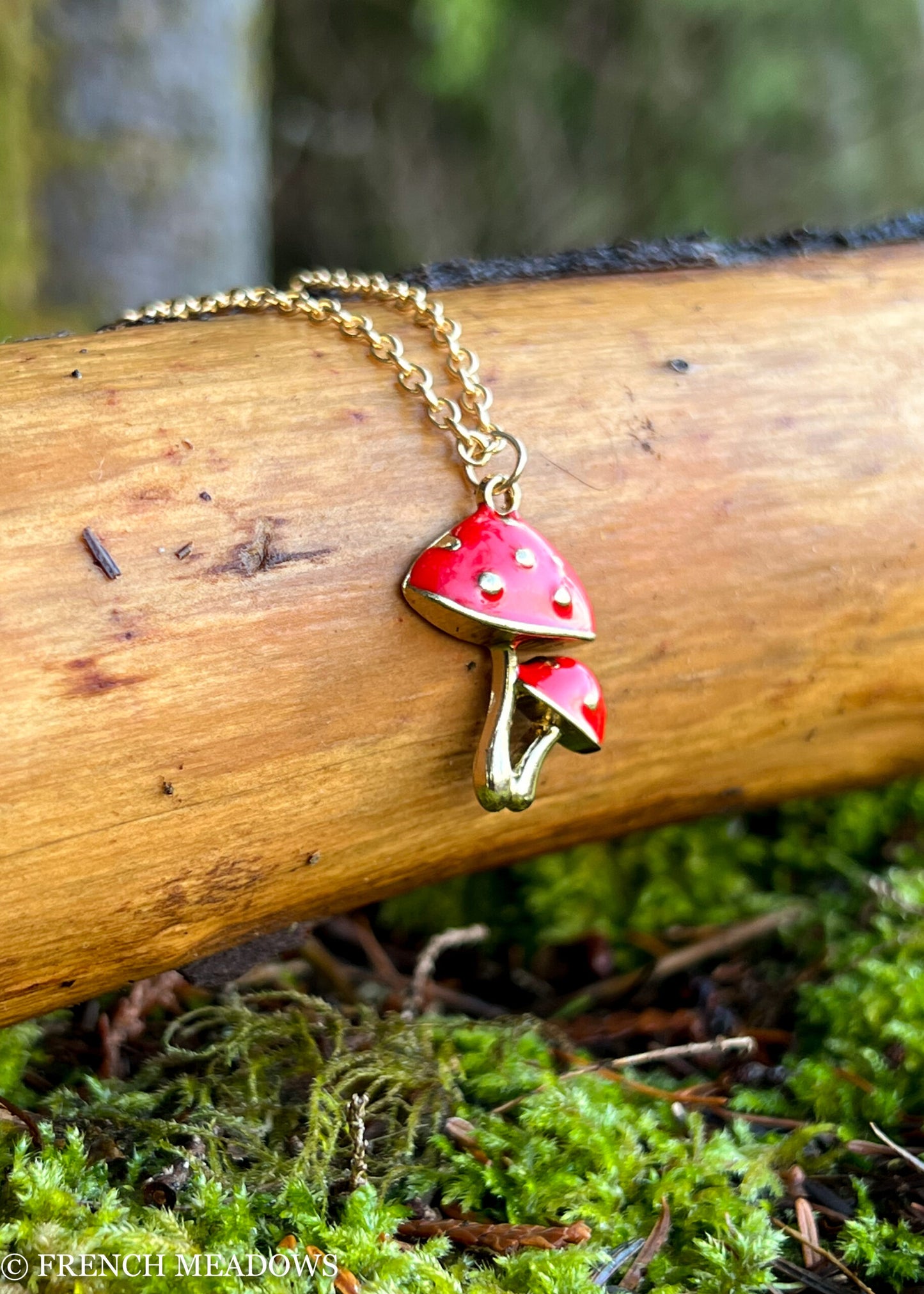 Amanita Mushroom Necklace with red and gold double mushroom pendant on gold chain, displayed on wooden log with mossy background