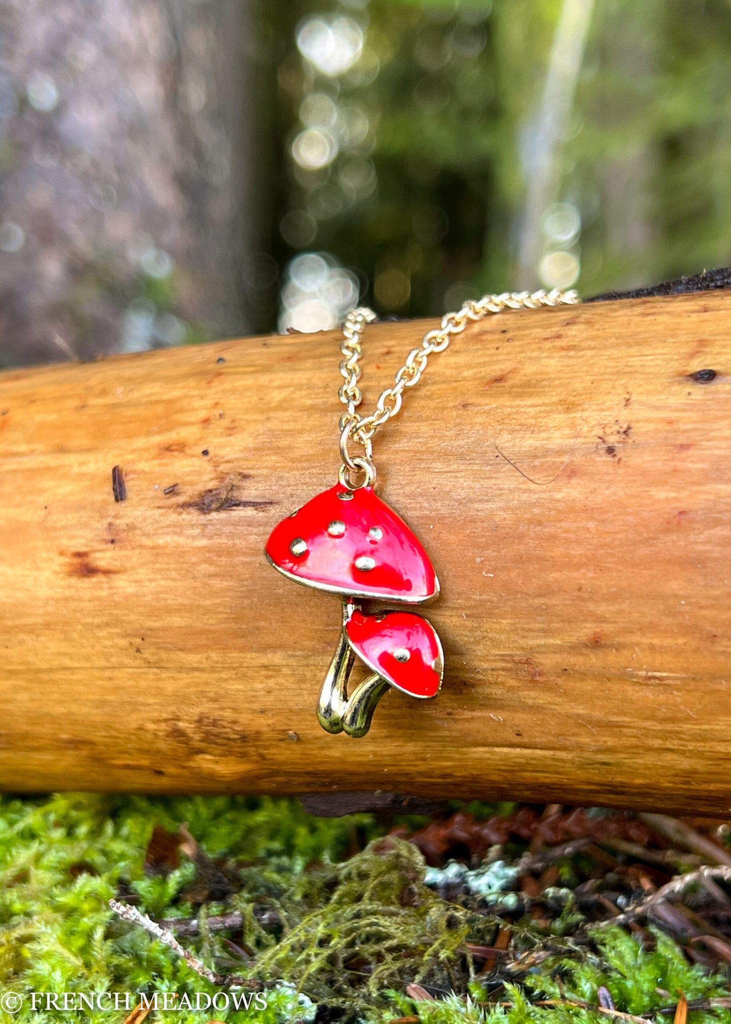 Amanita Mushroom Necklace with red spotted caps on gold chain, displayed on wooden log against natural forest backdrop