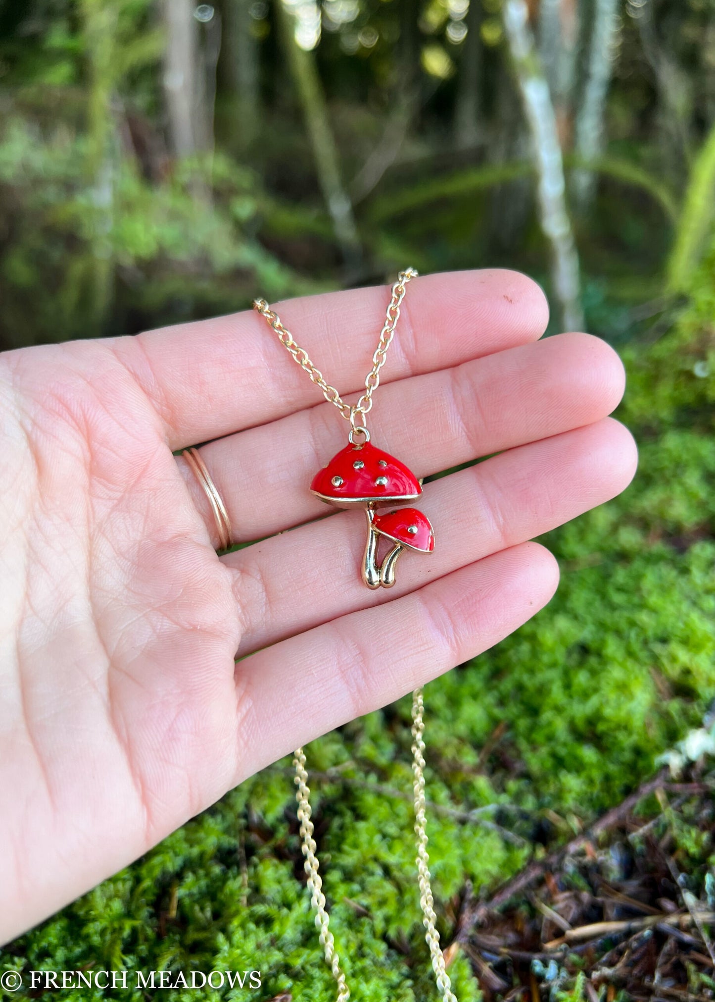Amanita Mushroom Necklace with red and gold double mushroom pendant displayed on palm against blurred forest background