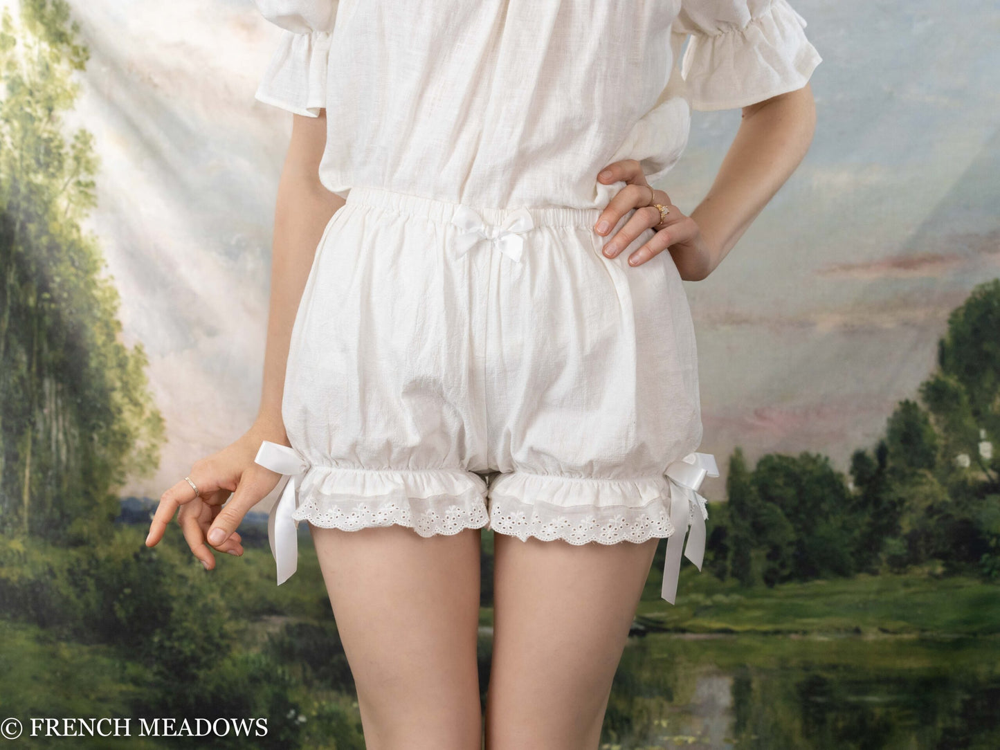 White Victorian Bloomers with lace trim and bow details, elastic waist and ruffled hem shown against pastoral background