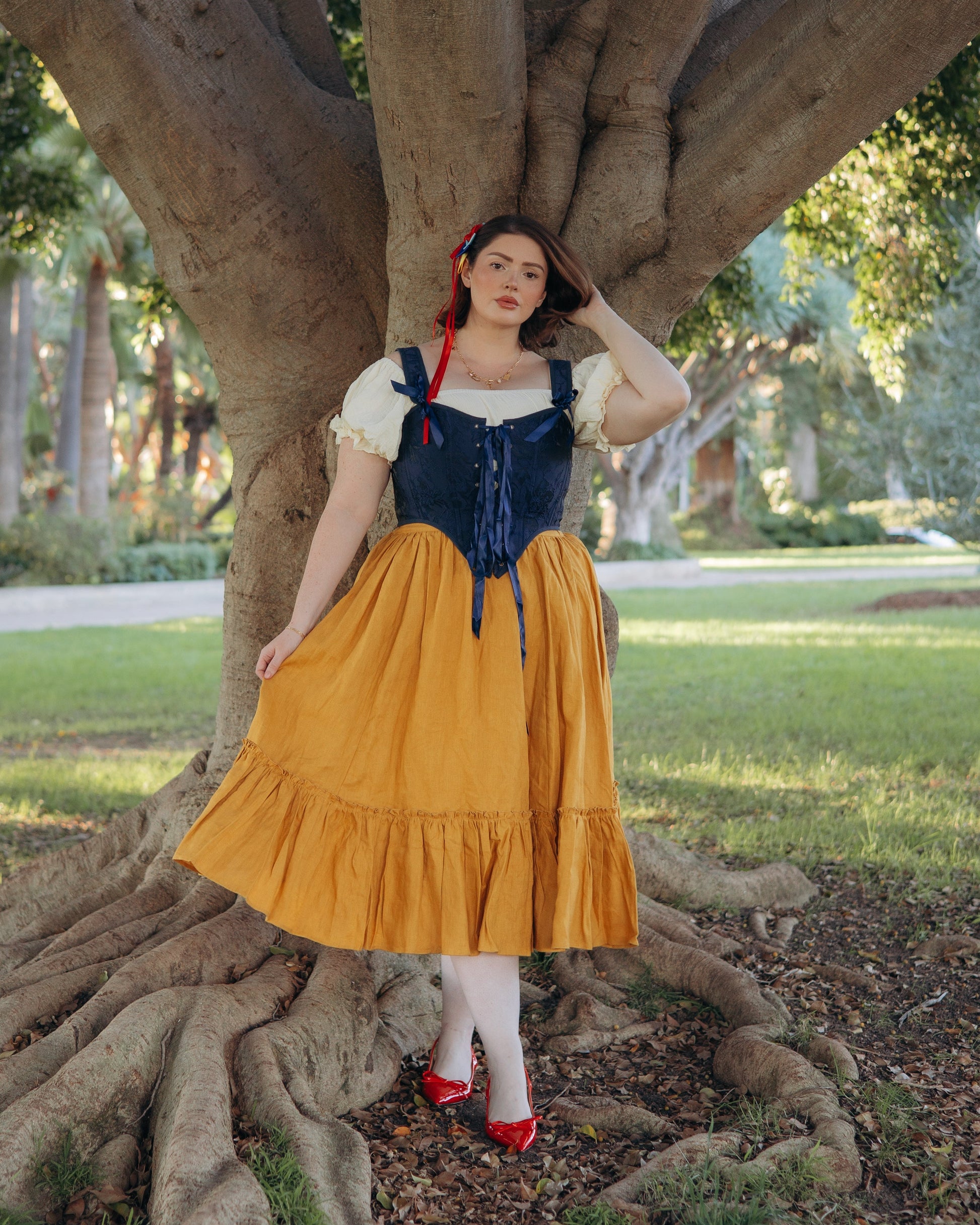 Woman wearing a dark blue floral jacquard corset paired with a yellow skirt standing by a large tree outdoors