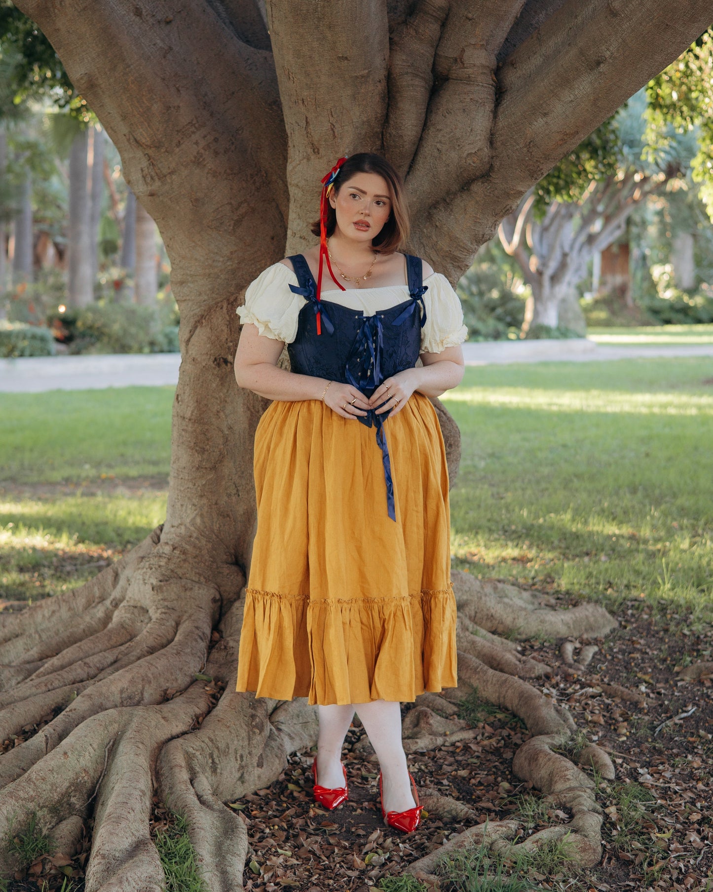 Woman wearing dark blue floral jacquard corset over white blouse with mustard skirt standing against tree roots outdoors