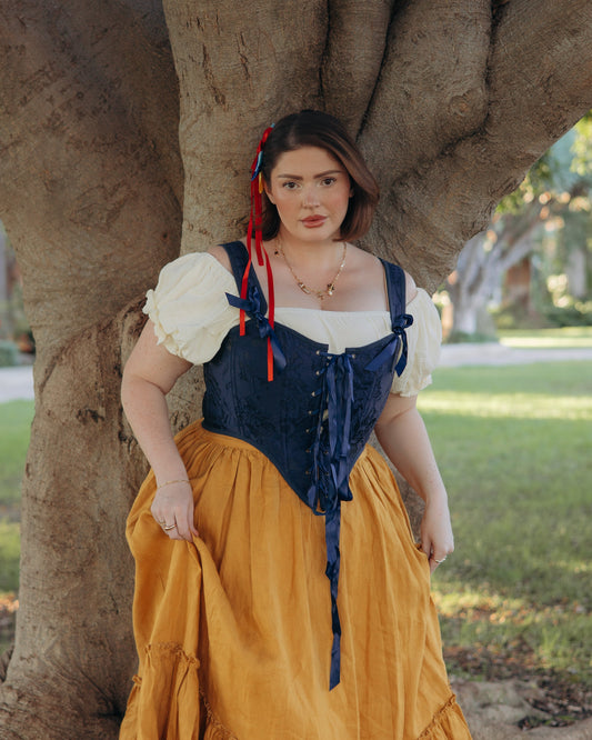Woman wearing dark blue floral jacquard corset with white blouse and mustard skirt standing by tree outdoors