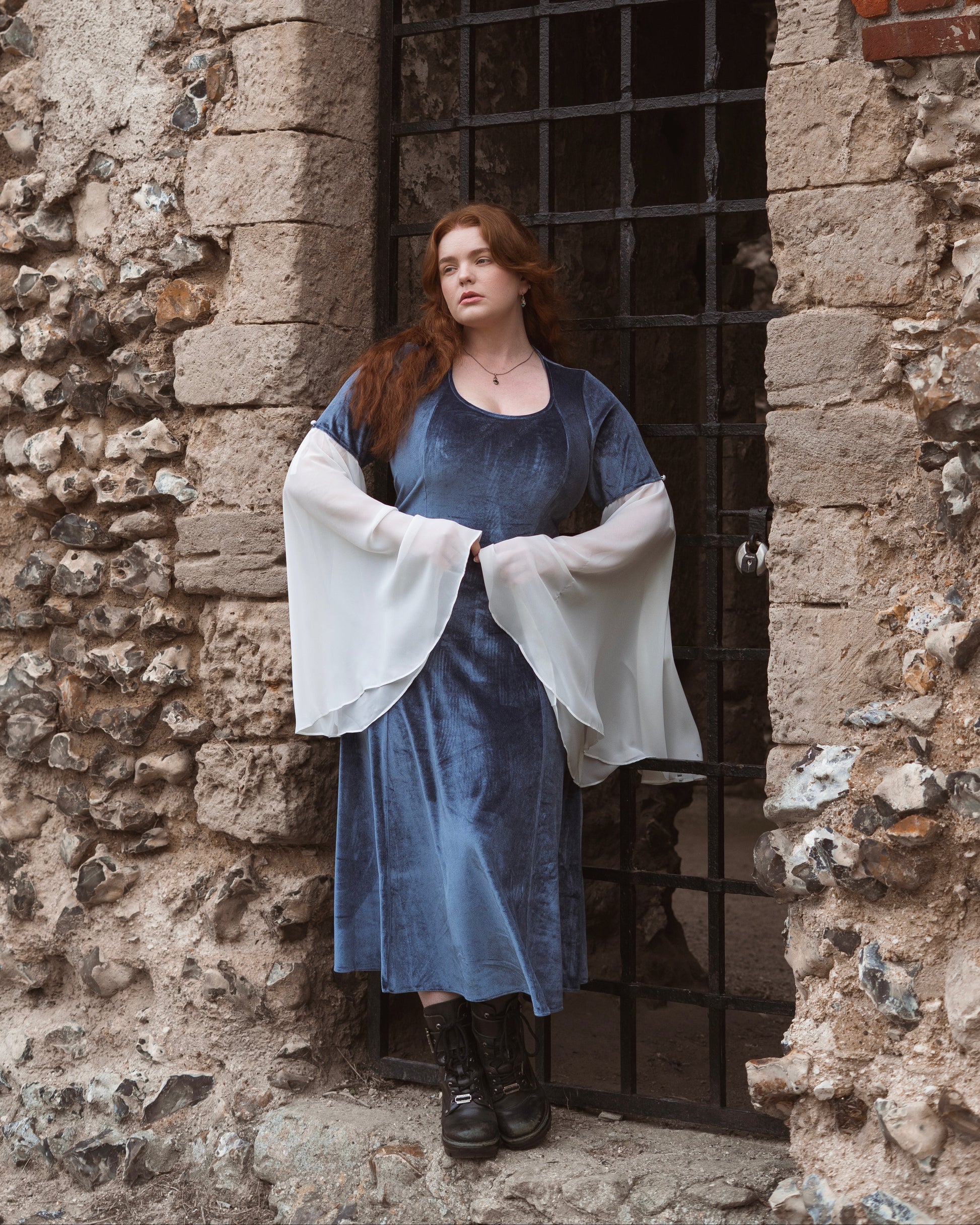 Woman wearing Elven Blue Velvet Bell Sleeve Dress with sheer white flowing sleeves standing by stone archway and metal gate