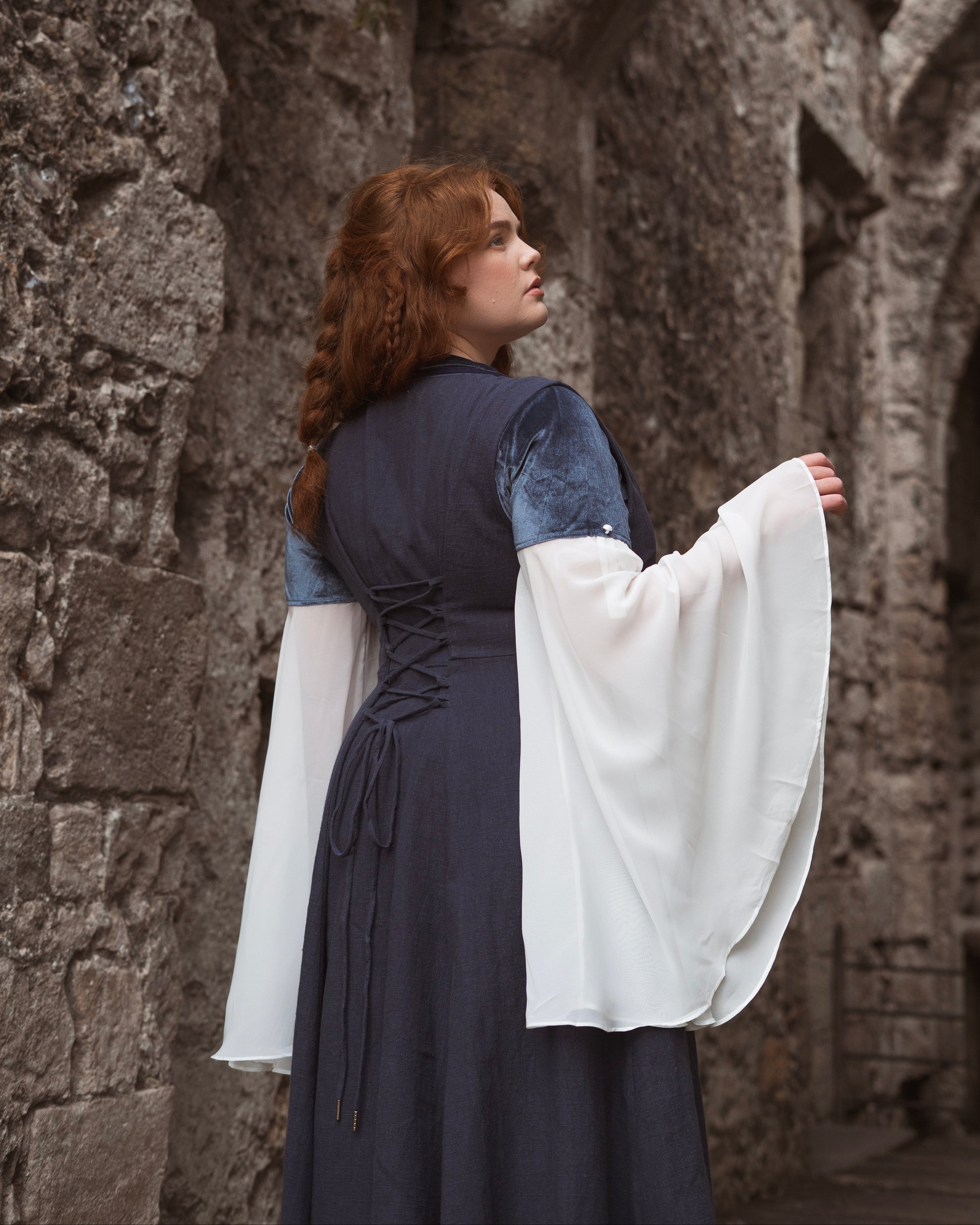 Woman wearing a navy blue Medieval-style kirtle dress with laced back and flowing white sleeves standing by stone walls