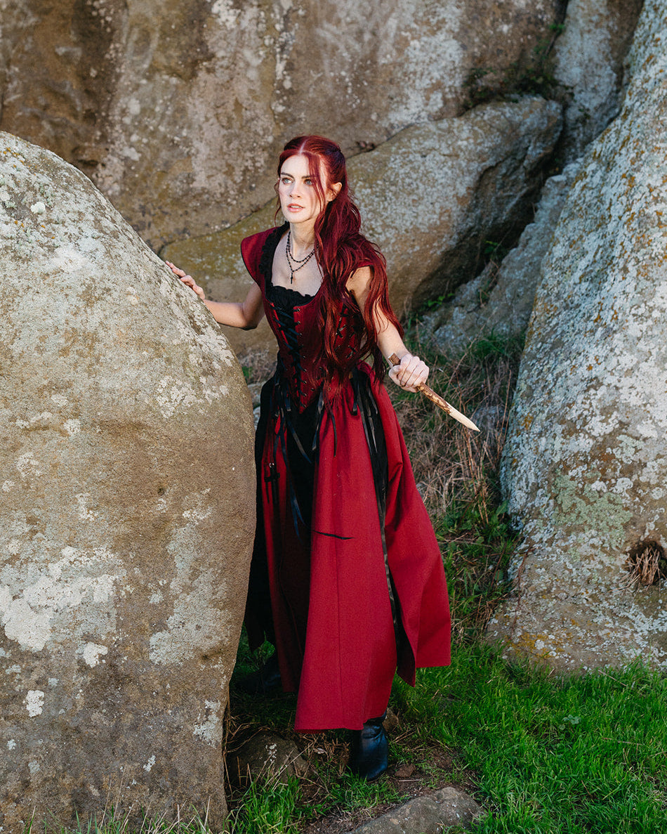 Woman with long red hair wearing a Red Vampire Slayer Corset Dress standing between large rocks outdoors holding a wooden dagger
