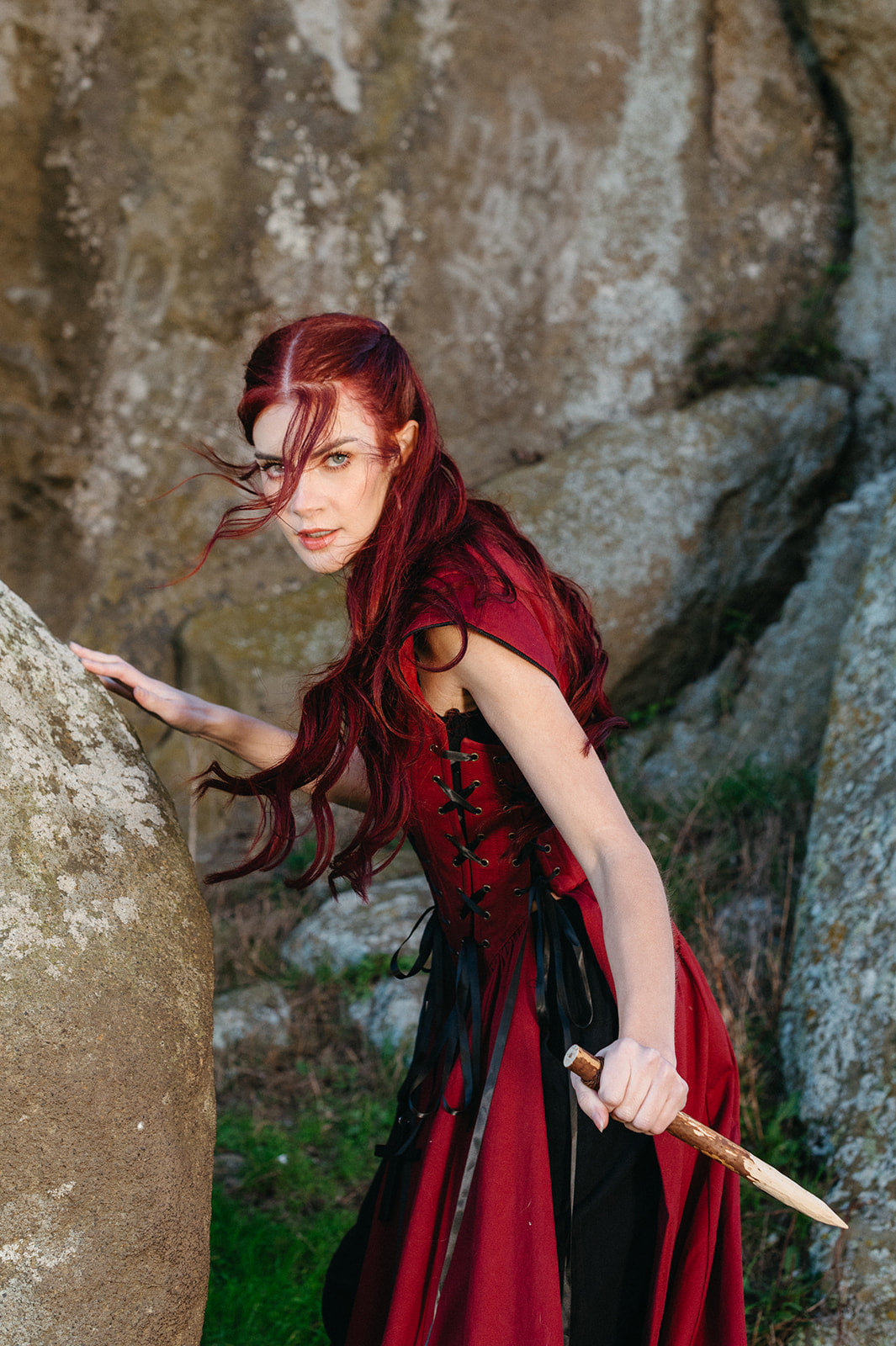 Woman with flowing red hair wielding a dagger wearing the Red Vampire Slayer Corset Dress outdoors by rocks