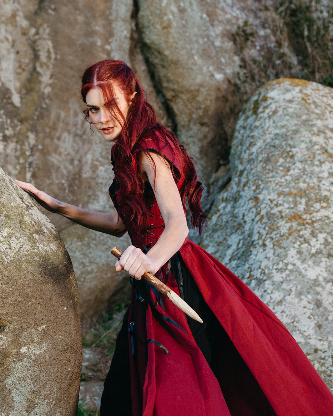 Woman with red hair wearing Red Vampire Slayer Corset Dress holding wooden stake near large rocks outdoors