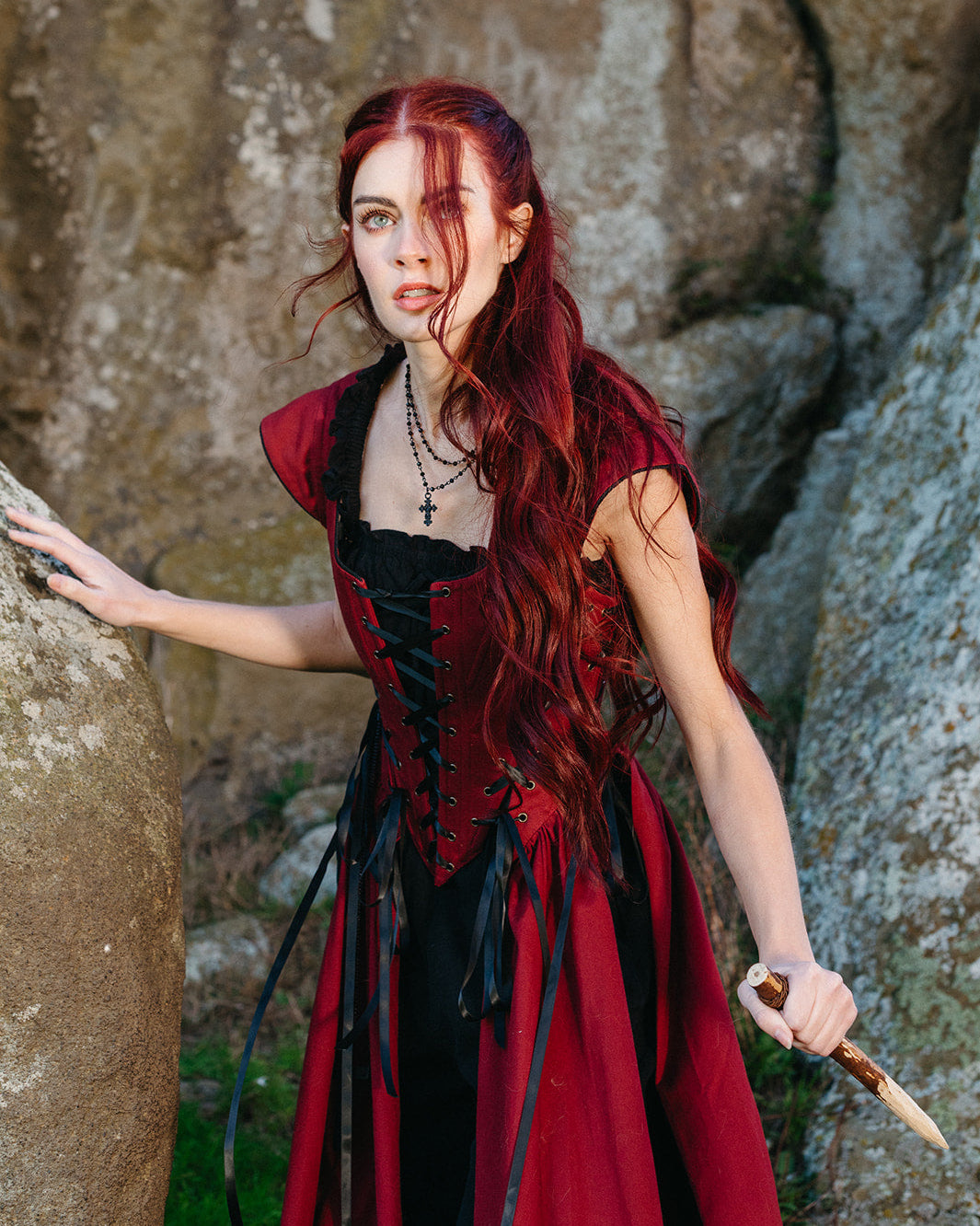 Woman in a Red Vampire Slayer Corset Dress with long red hair holding a dagger poised in rocky outdoor setting