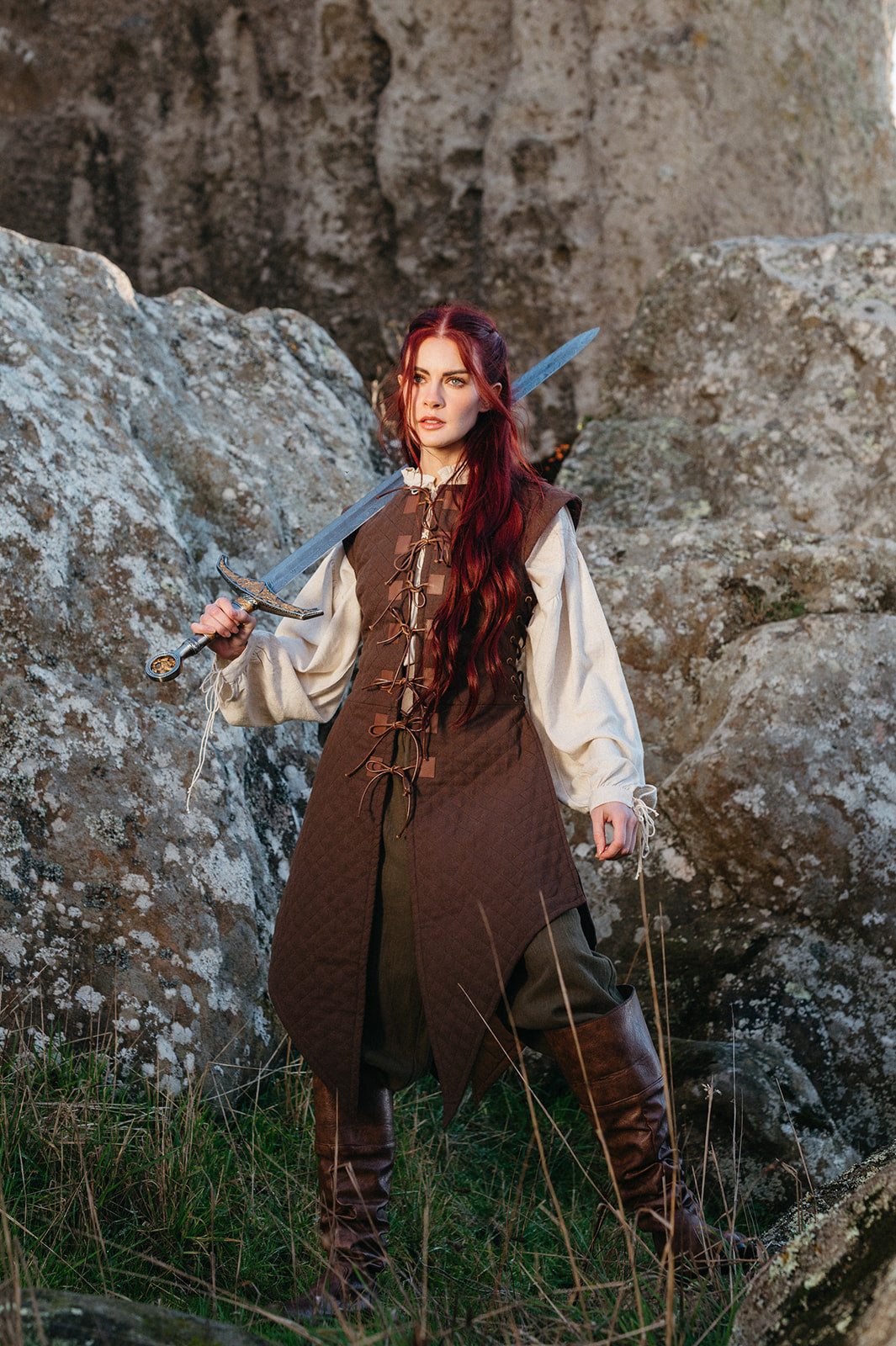 Woman with long red hair wearing a Quilted Gambeson holding a sword standing among rocky terrain outdoors
