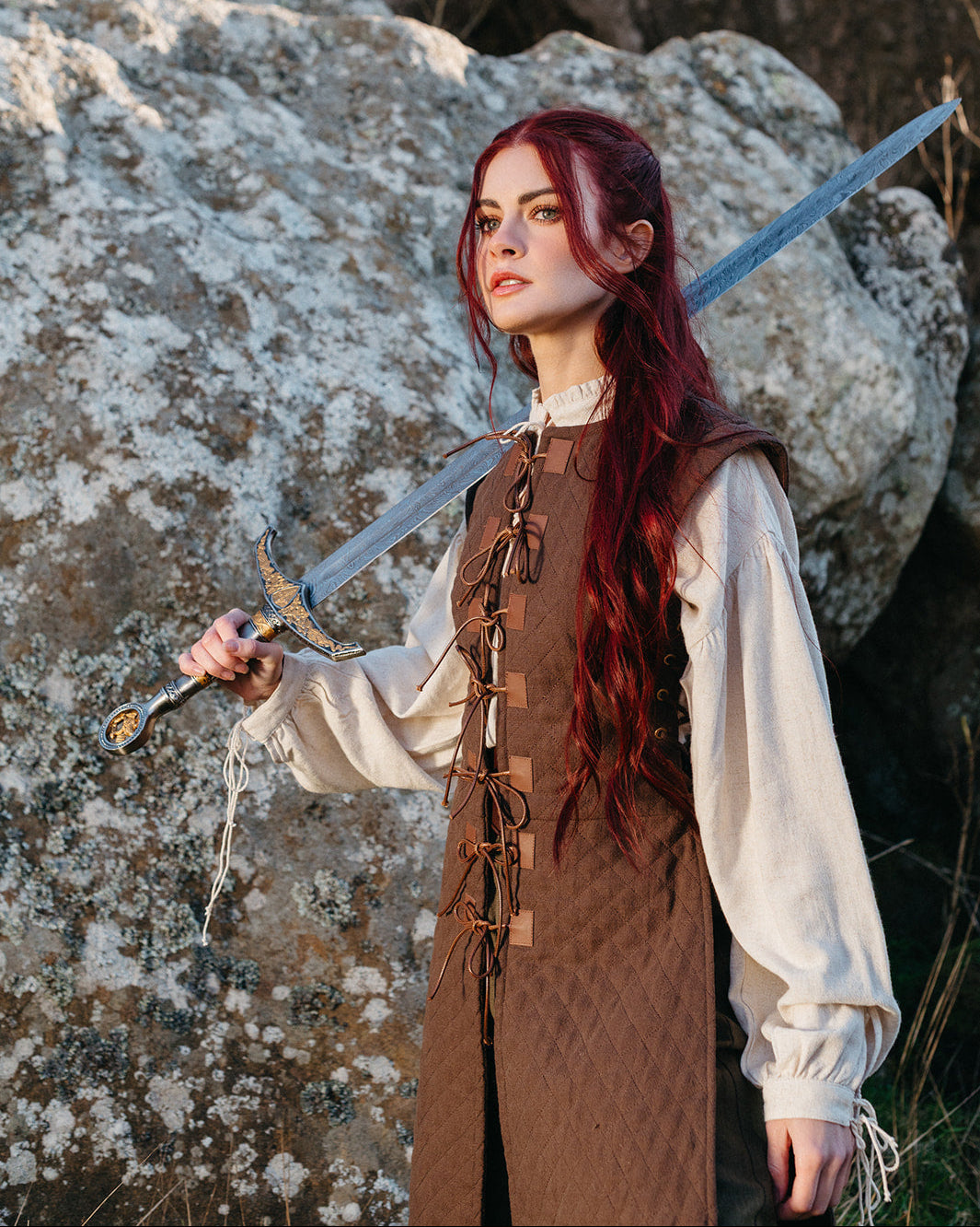 Woman wearing a brown Quilted Gambeson with long sleeves holding a medieval sword outdoors by rocky terrain