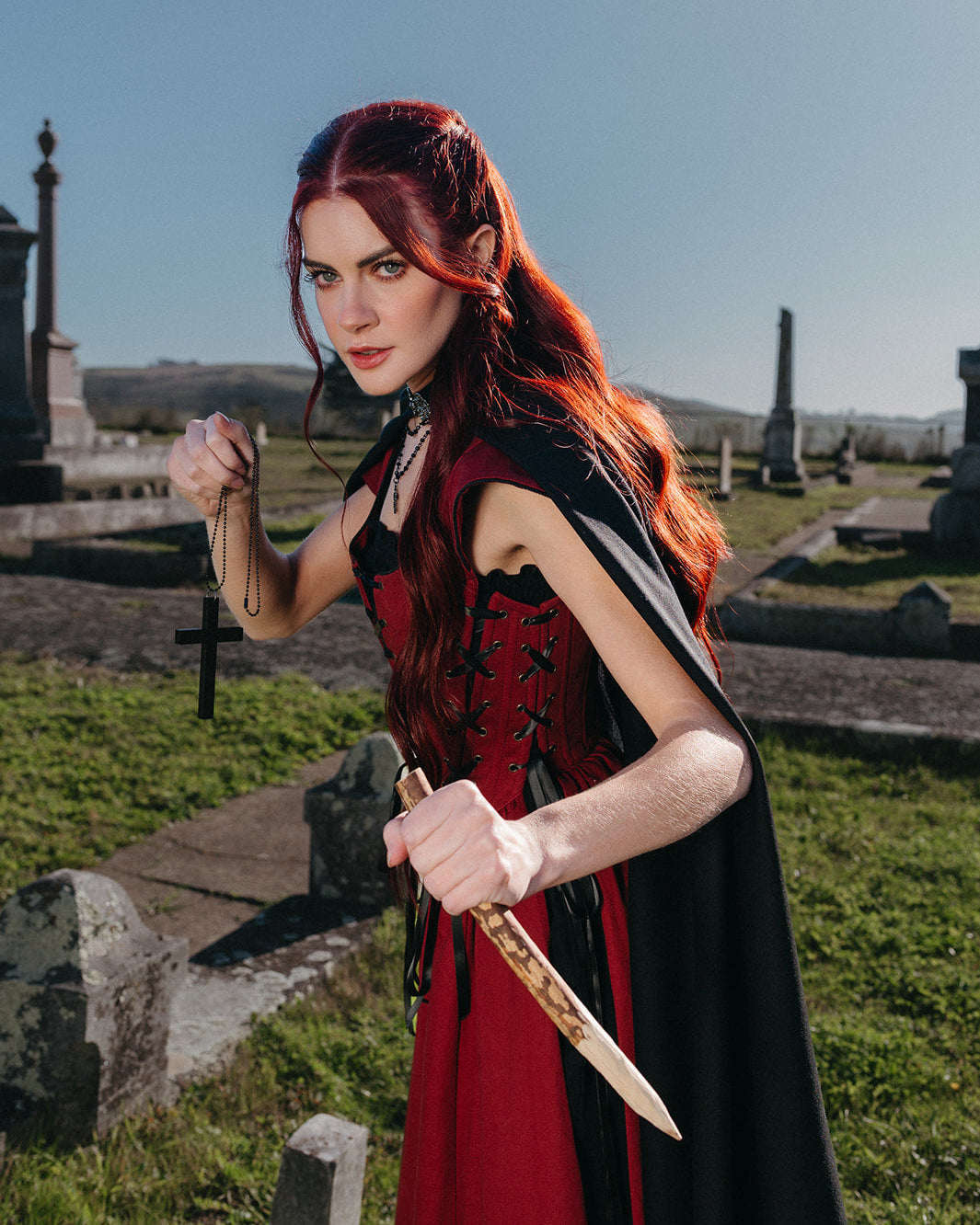 Woman in graveyard wearing Red Vampire Slayer Corset Dress holding wooden stake and cross necklace ready to defend