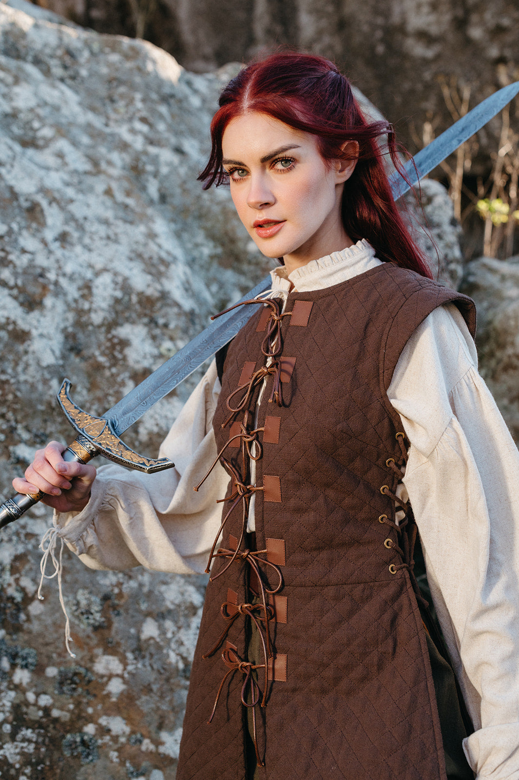 Woman wearing a brown Quilted Gambeson holding a medieval sword with rocky background outdoors
