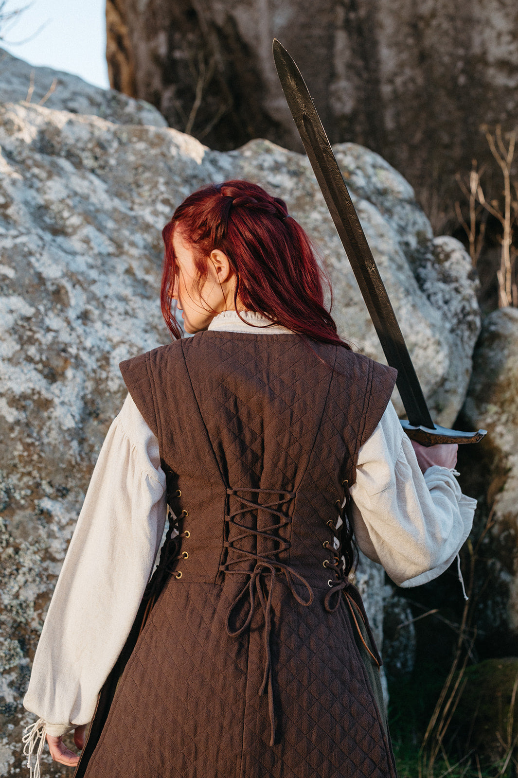 Woman holding sword outdoors wearing brown quilted gambeson with lacing details on back and white sleeves