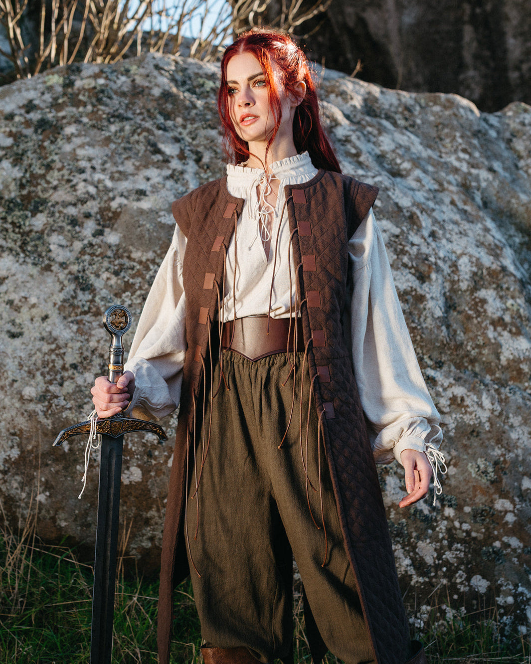 Woman wearing a brown Quilted Gambeson over medieval blouse and pants holding a sword in a natural rocky outdoor setting.