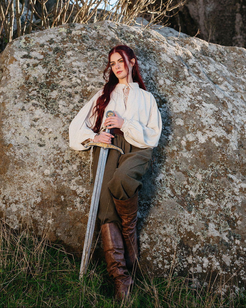 Woman wearing Borgia Shirt holding a sword while leaning against a large rock in a natural outdoor setting