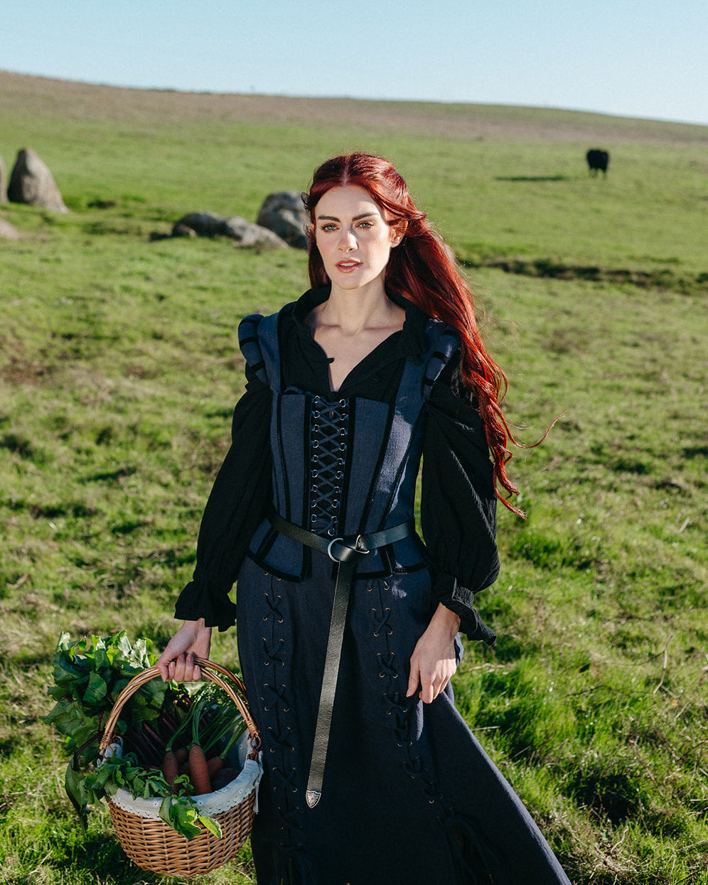 Woman wearing Navy Tavern Bodice with laced front and belt, standing in a grassy field holding a basket of vegetables