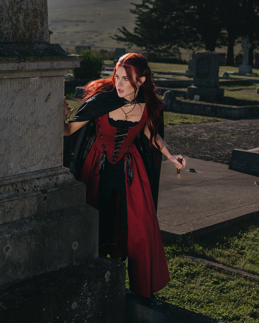Woman in a dark graveyard wearing a red and black Red Vampire Slayer Corset Dress with lacing and cape details