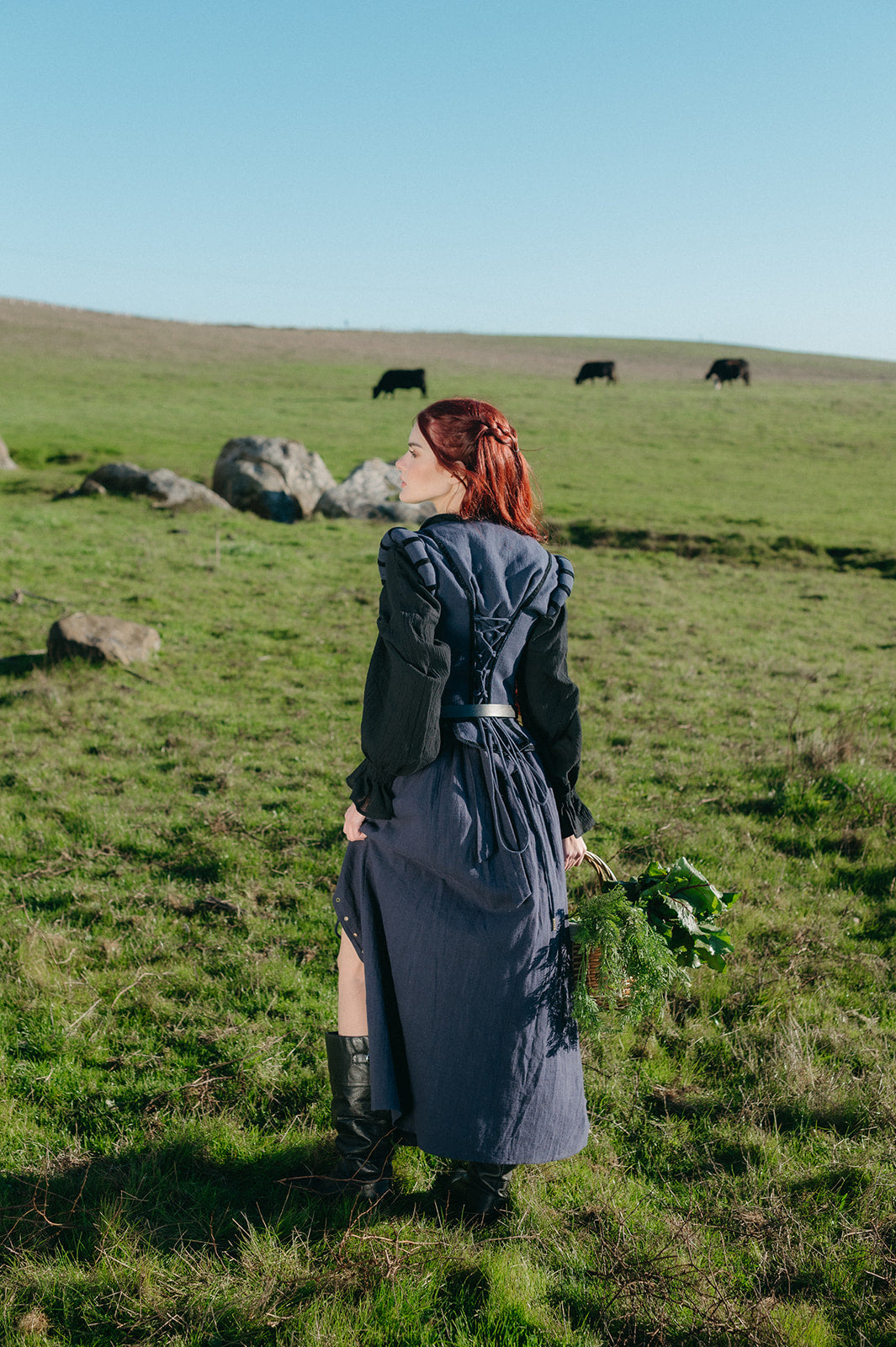 woman wearing navy tavern bodice standing in grassy field holding fresh greens under clear blue sky