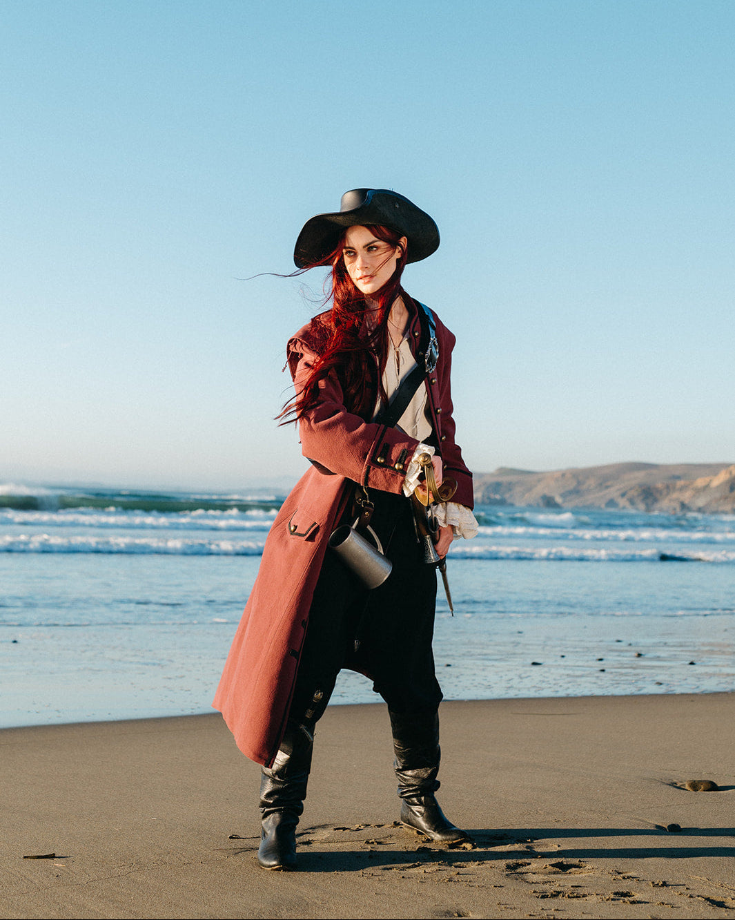 Woman wearing a Pirate Captain's Coat standing on a beach with ocean waves and hills in the background