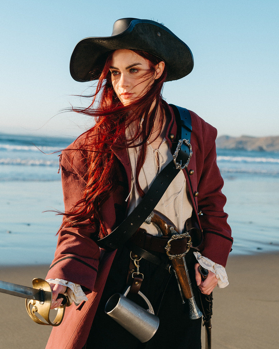 Woman on beach wearing Leather Pirate Belt with sword, vintage jacket, and wide-brimmed hat in dramatic seaside scene