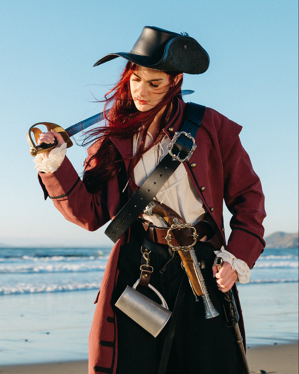 Woman wearing Pirate Captain's Coat holding a sword and flintlock pistol standing on a beach at sunset