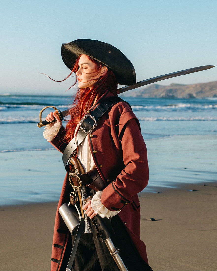 Woman wearing a Pirate Captain's Coat with a tricorne hat holding a sword on a beach at sunset
