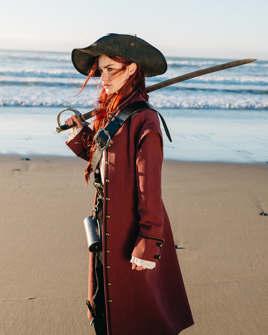 Woman wearing Lacey Ruffle Sleeves peeking from under a burgundy coat holding a sword by the beach at sunset