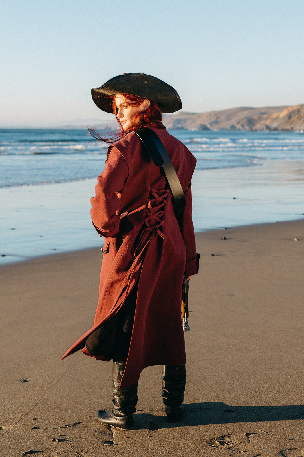 Woman wearing the Pirate Captain's Coat with lace-up back detail standing on a beach at sunset