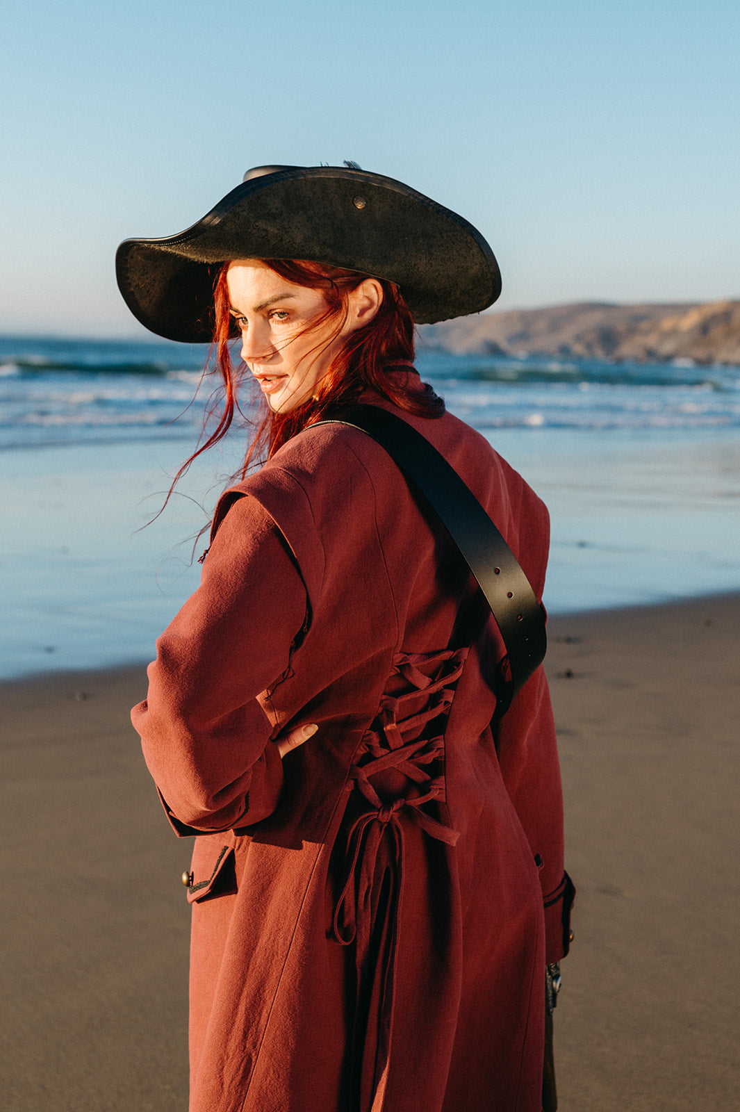 Woman wearing a rust-colored Pirate Captain's Coat with laced back detail and black tricorn hat on the beach at sunset