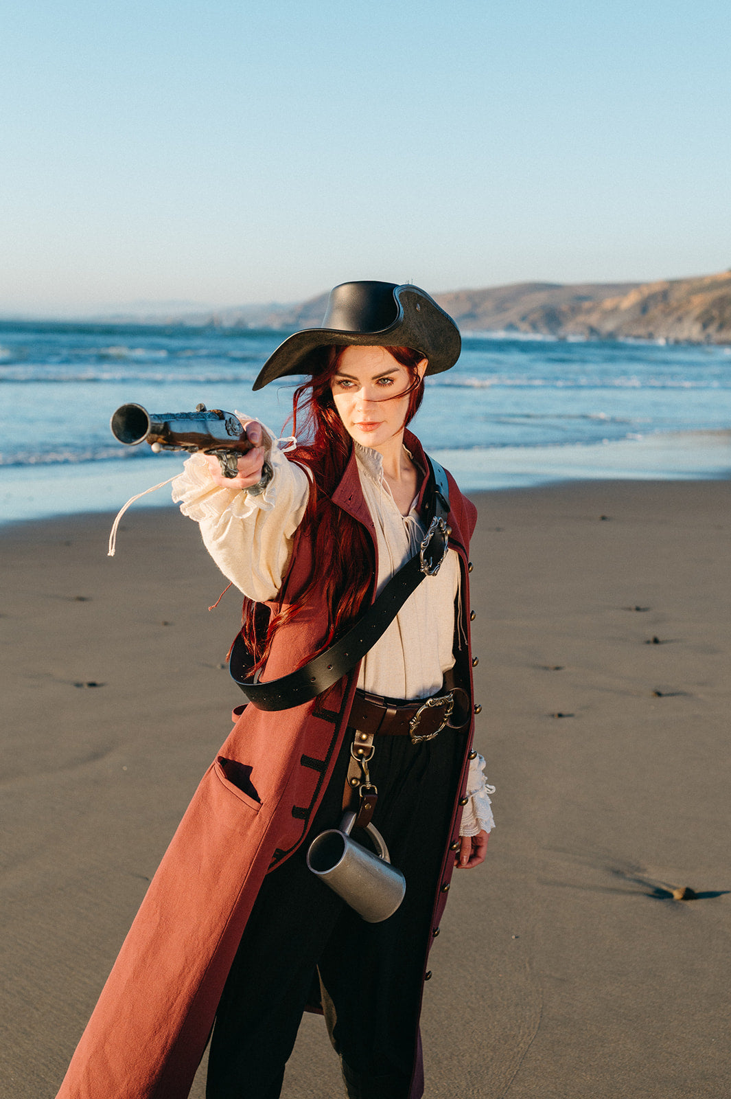 Woman wearing Pirate Captain's Coat and tricorn hat pointing pistol on a sandy beach with ocean background