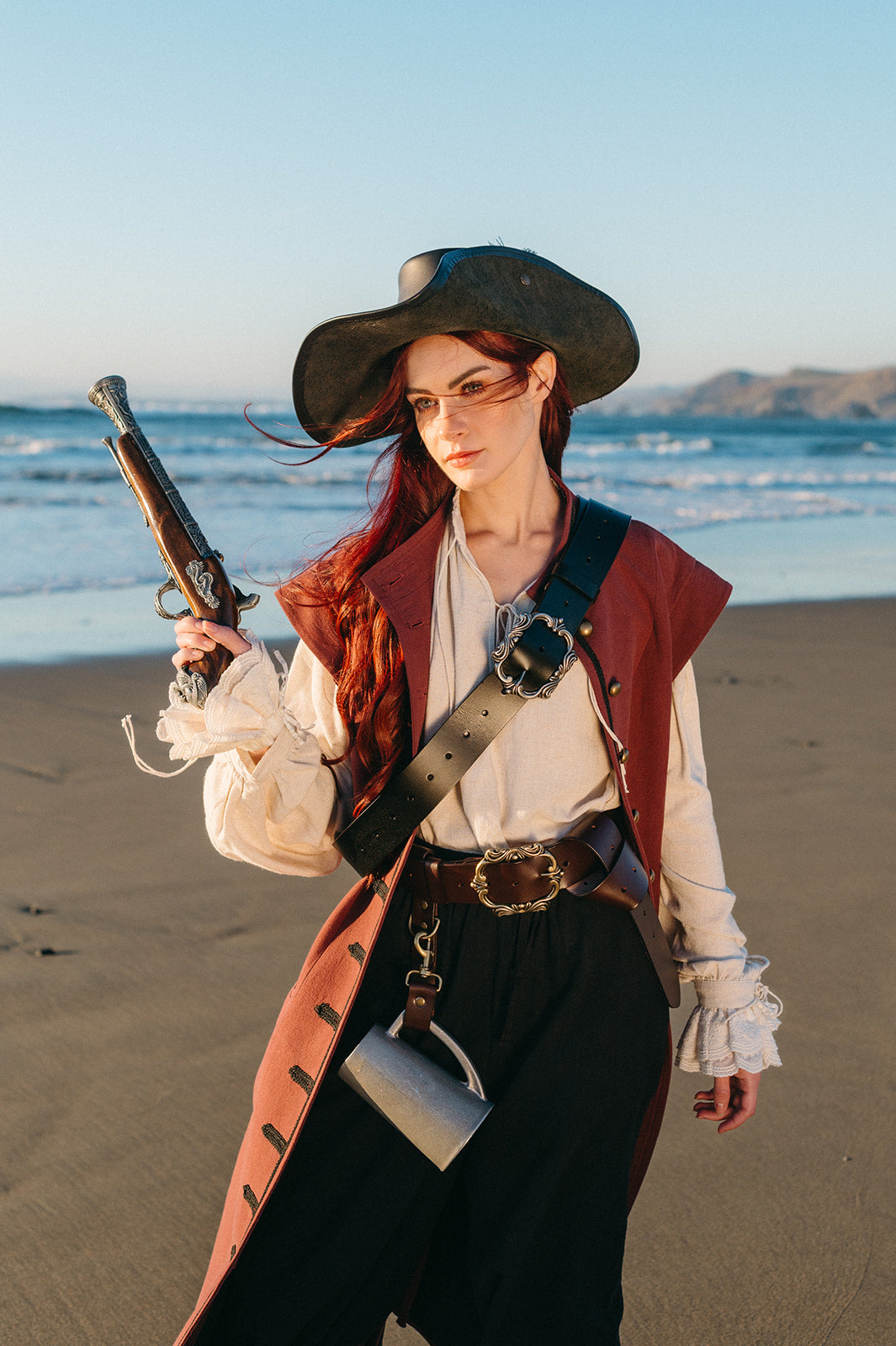 Red-haired woman in a burgundy vest and cream blouse wearing a wide-brimmed hat holding a vintage pistol on the beach