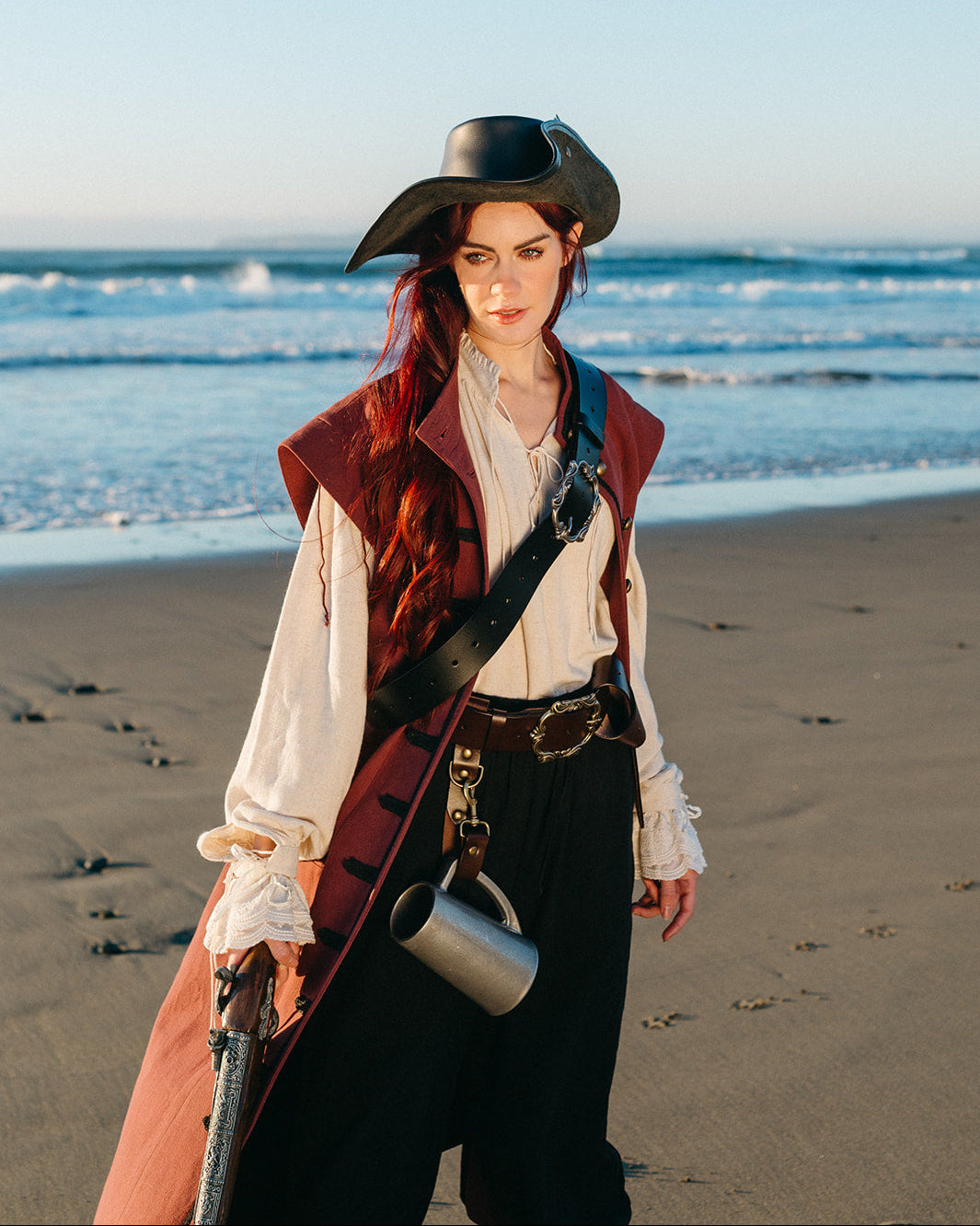 Woman wearing a Pirate Captain's Coat with a tricorn hat on a beach holding a vintage flintlock pistol
