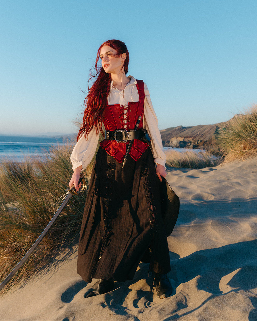 Woman standing on beach wearing a Leather Pirate Belt and holding a sword with ocean and dunes in the background