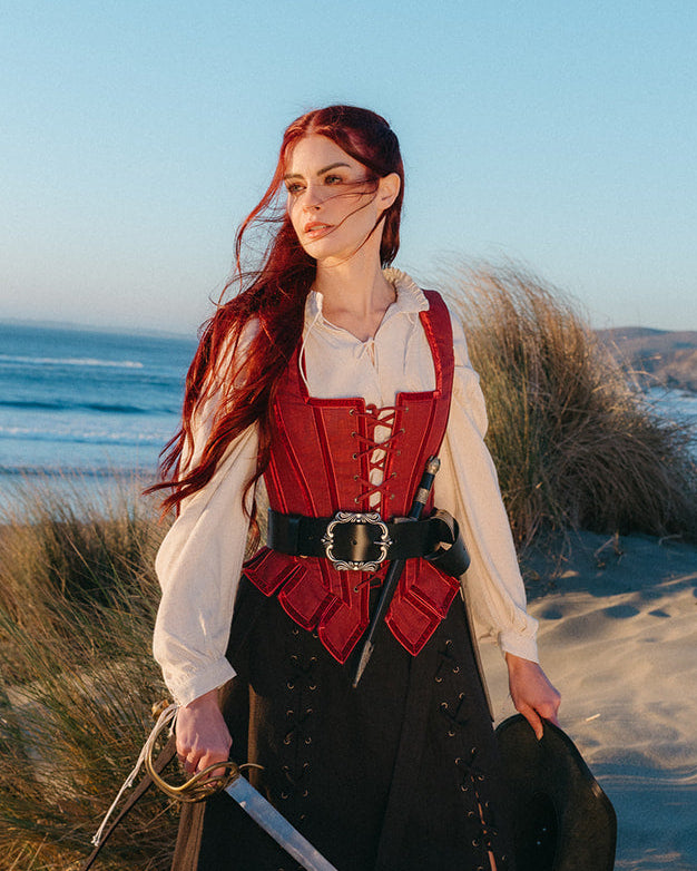 Woman standing on beach wearing a Red Pirate Doublet with a black belt and holding a sword and hat in hand